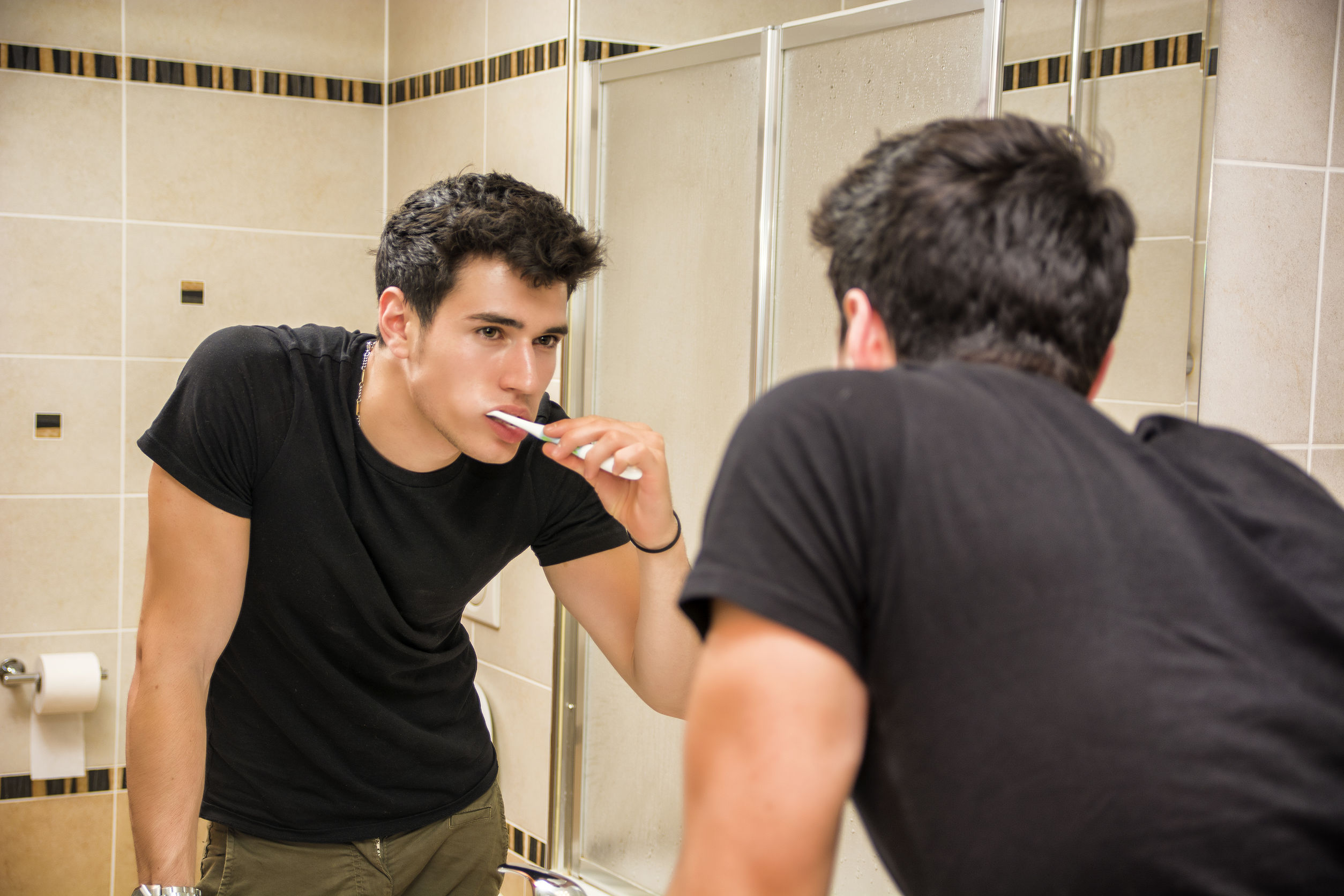 Headshot-of-attractive-young-man-brushing-teeth-with-toothbrush-looking-at-himself-in-mirror