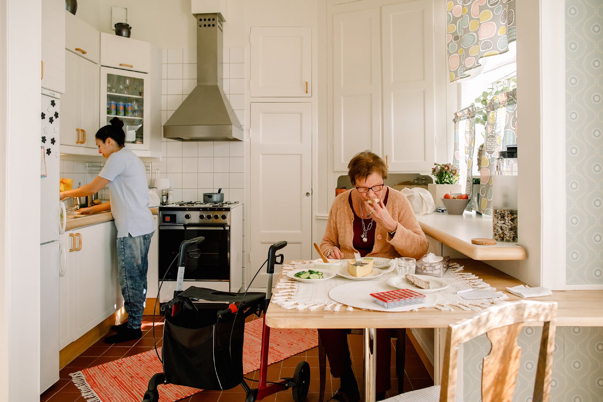 Special Benefits Insurance Services | Planning On Independent Living? Health Insurance Can Help With Healthcare Costs Old woman with an assistive walking device eating at the kitchen table, while an assistant works at the kitchen counter