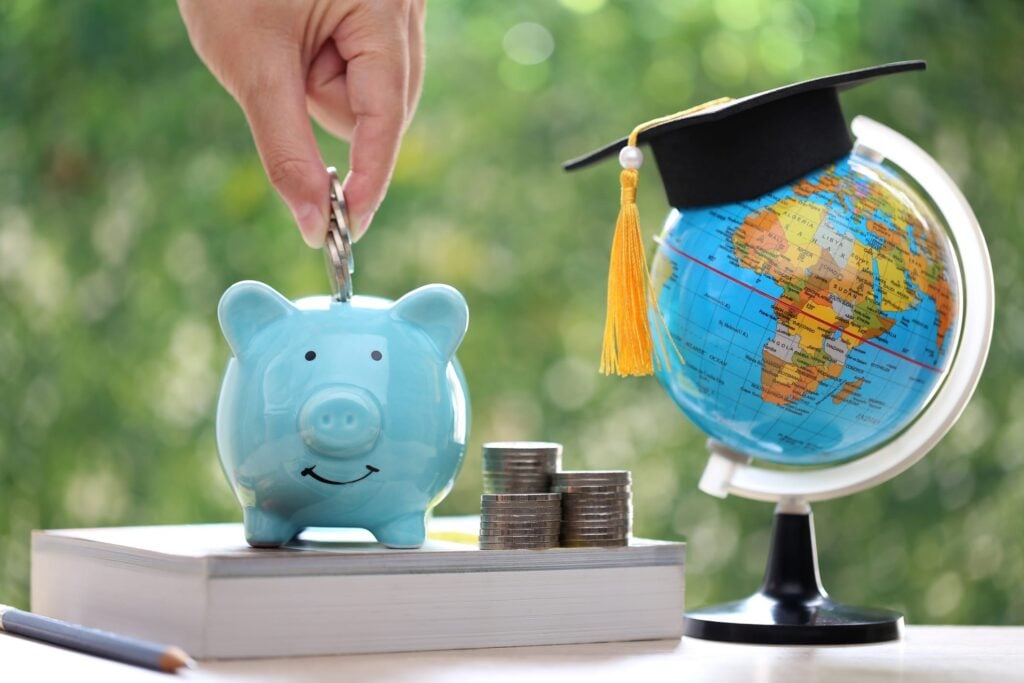 Hand inserting coins into a blue piggy bank, next to a globe with a graduation cap