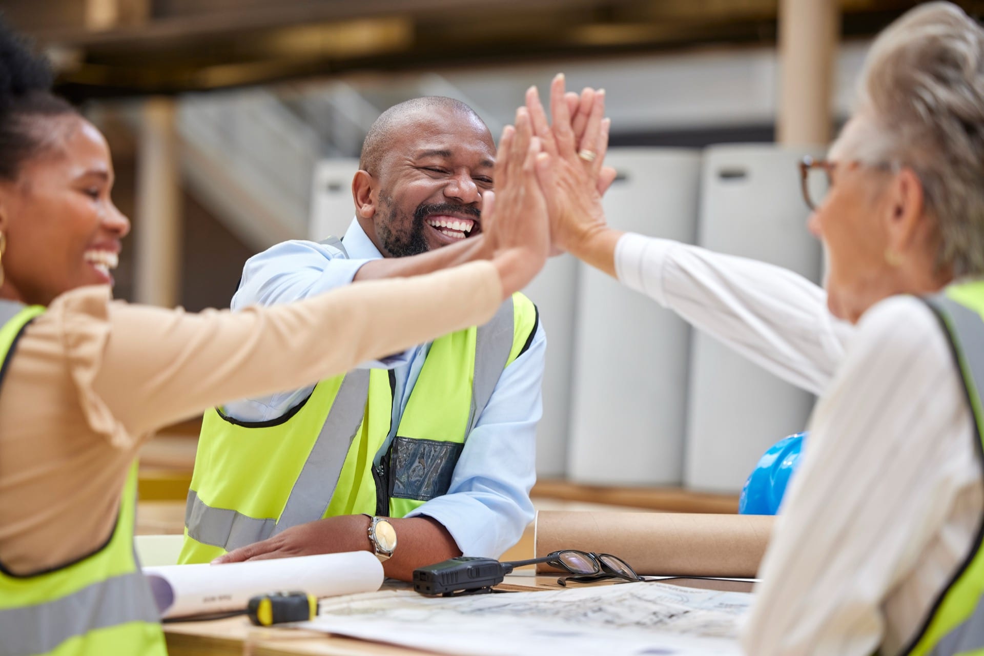 A diverse team of smiling workers in safety vests giving a group high-five, celebrating successful teamwork in an industrial setting.