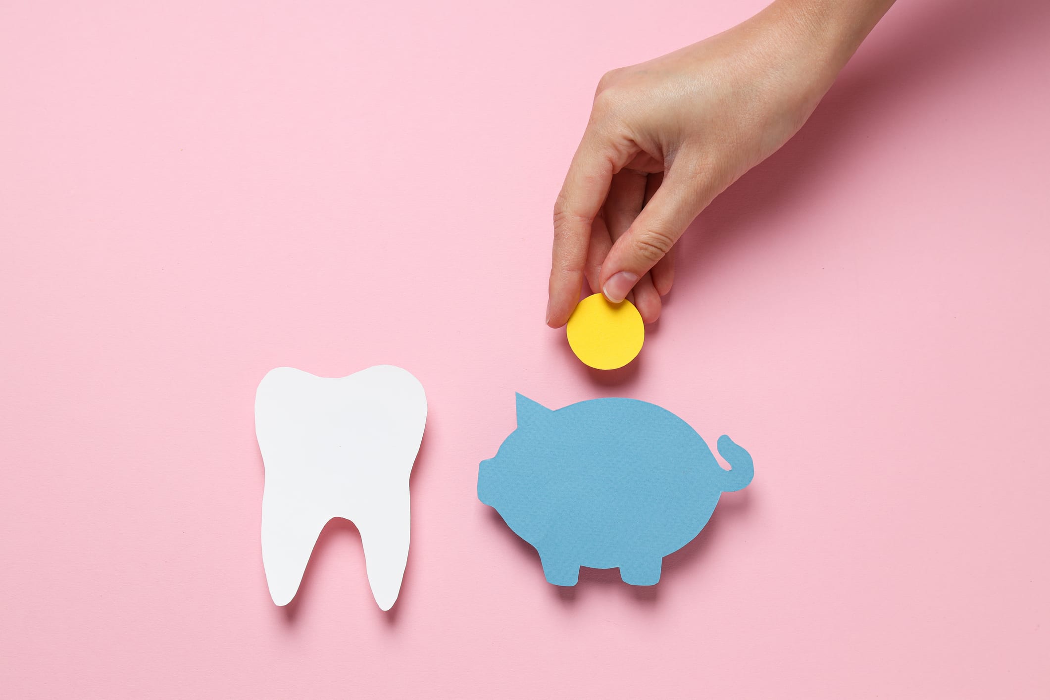 Special Benefits Insurance Services | 5 CDCP Hidden Secrets And Why Dental Insurance Still Wins Hand dropping a yellow coin into a blue paper cutout of a piggy bank next to a white paper cutout of a tooth, symbolizing dental savings or insurance costs.
