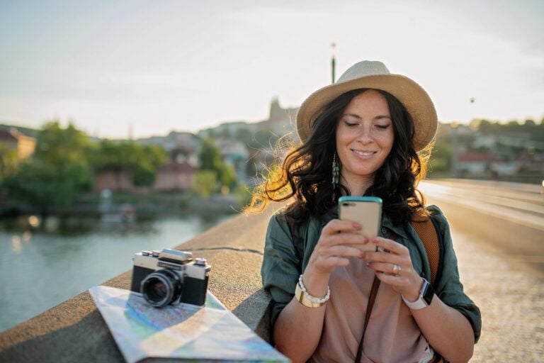 Smiling female tourist in sunhat using a smartphone on a city bridge with a map and vintage camera nearby, illustrating a travel planning app.